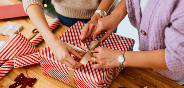 Escena de dos mujeres envolviendo un regalo para el Día de la Madre, transmiten cercanía, detalle y el cariño de preparar un obsequio especial en esta fecha.