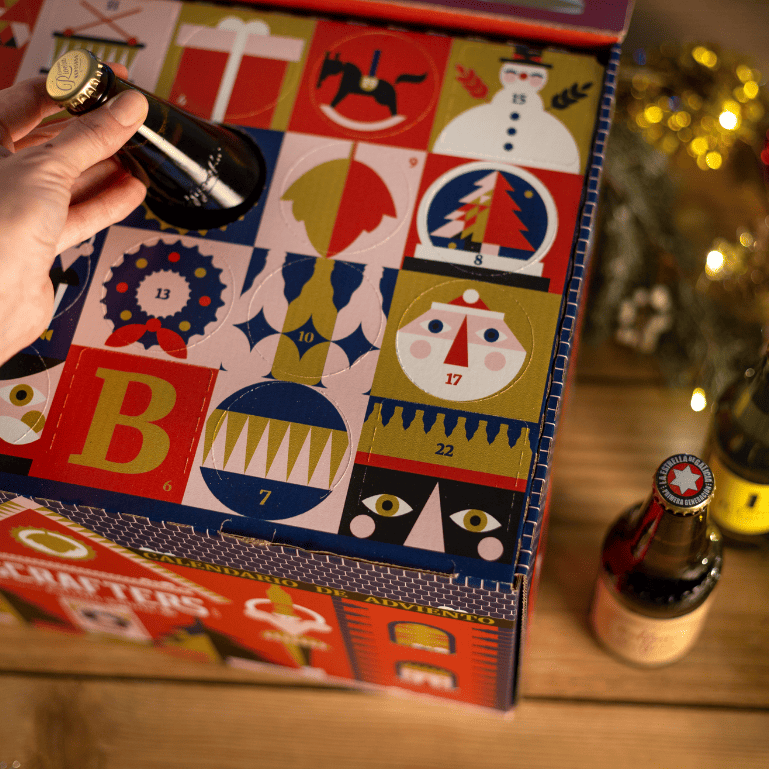 Una persona saca una botella de Rivera Reposada del Calendario de Adviento de cervezas Bigcrafters en caja con forma de casita navideña ilustrada por Yarza Twins.