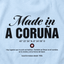 Primer plano de tela de algodón azul claro de camiseta con serigrafía negra detallada del texto "Hecho en A Coruña", coordenadas, logotipo de estrella y una cita en español.