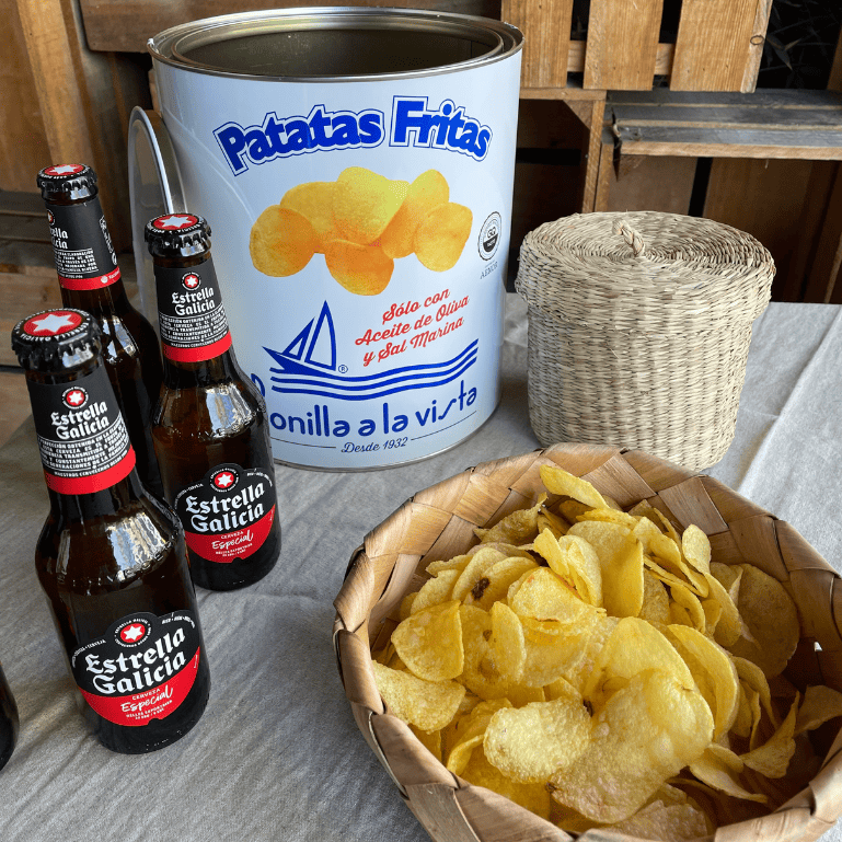 Cervezas Estrella Galicia Especial de 25 cl en una mesa, con su aroma maltoso y carácter refrescante, se complementa con una lata de patatas Bonilla a la vista de 500 g que aporta un crujido salado y textura ligera.  