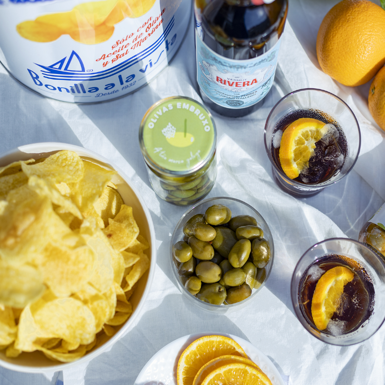 Dos vasos y una botella de Vermut Rivera acompañado con olivas verdes y patatas Fritas de Bonilla A La Vista, que combinan bien sus sabores y texturas para ofrecer un aperitivo delicioso