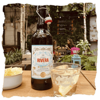 una vaso y una botella de Vermut Rivera blanco, clásico con lúpulo Nugget gallego, aroma herbacéo y notas de genciana, corteza de naranja y ruibarbo, ofreciendo un sabor moderado y amargo