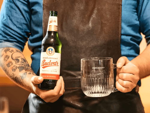 Un hombre sostiene una jarra y una botella de cerveza checa Budvar, que es refrescante, con una espuma densa y un sabor malteado que la convierte en la cerveza perfecta para acompañar comidas tradicionales del centro de Europa