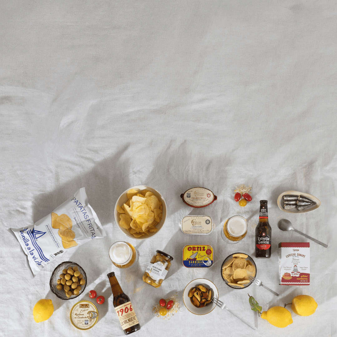 Packs de aperitivos con cervezas Estrella Galicia y 1906, patatas Bonilla a la Vista, conservas Ortiz, aceitunas, mejillones y pescado ahumado sobre mantel de color blanco.